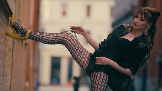Model in Ellipse Net fishnet tights and yellow heels kicking one leg up against a brick wall, stretching the net with her hand